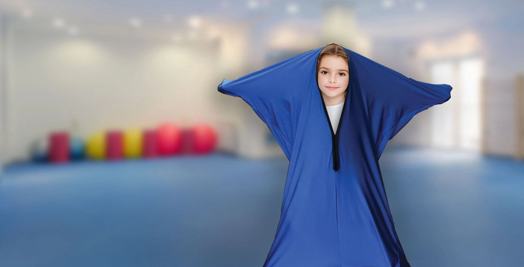 Child wearing a blue sensory body sock in an indoor therapy setting with colorful exercise balls in the background, demonstrating compression and calming use.