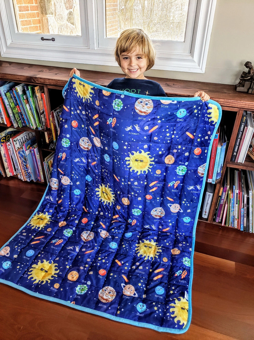 Child smiling while holding a Solar System weighted blanket featuring planets and stars. This 5 lb sensory comfort blanket promotes calm, focus, and better sleep for children dealing with autism or sensory challenges. Soft, cozy, and educational — part of the Zula Autism Mart sensory comfort collection.